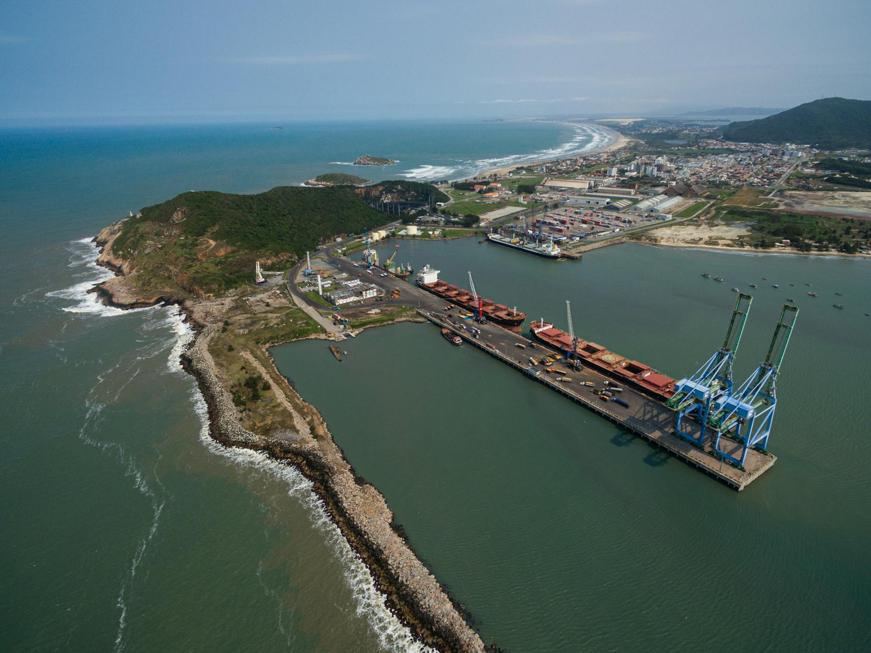 Vista aérea de um porto com navios, cais e guindastes, ao lado de costa e cidade.